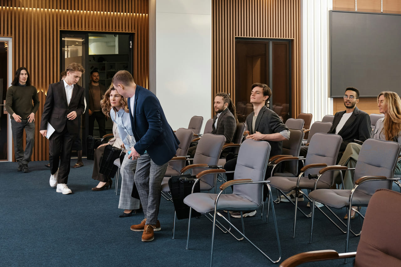 People arriving for a session in a conference room with wooden wall slats: a man carrying a folder walks in, others lean in to choose where to sit, and some are already seated facing the front.